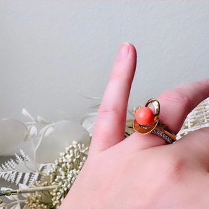 This vintage faux coral and gold Avon ring is a pretty little find. Gold tone ring featuring a vibrant faux coral stone in the center of an intricate gold arch loop. Major abstract space vibes! A unique statement piece by Avon from 1975. Signed in excellent vintage condition. 

Dimensions: US size 6-7 has inner sizer for flexible fit