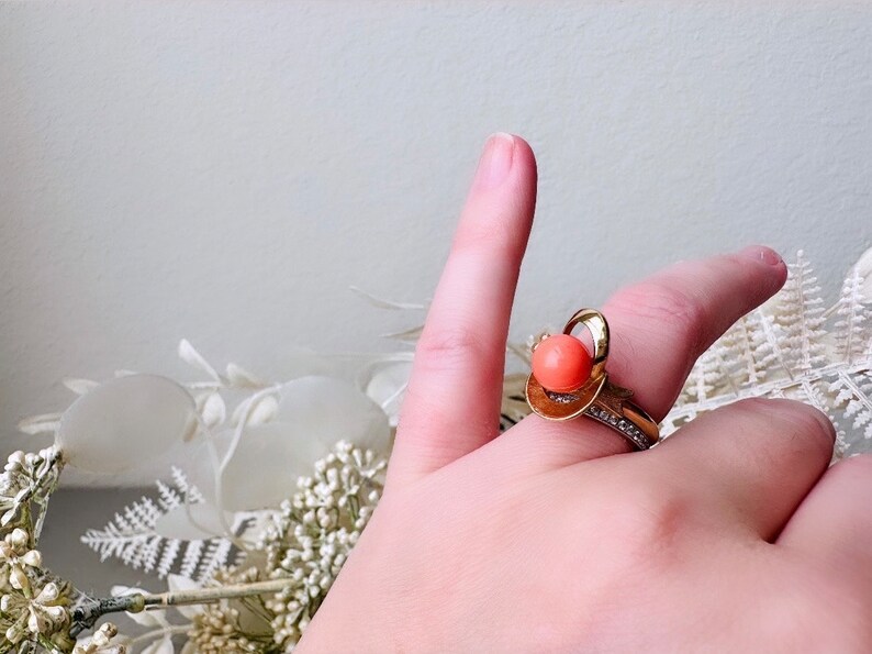 This vintage faux coral and gold Avon ring is a pretty little find. Gold tone ring featuring a vibrant faux coral stone in the center of an intricate gold arch loop. Major abstract space vibes! A unique statement piece by Avon from 1975. Signed in excellent vintage condition. 

Dimensions: US size 6-7 has inner sizer for flexible fit