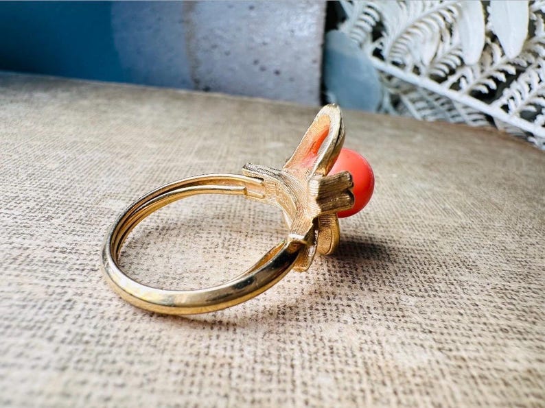 This vintage faux coral and gold Avon ring is a pretty little find. Gold tone ring featuring a vibrant faux coral stone in the center of an intricate gold arch loop. Major abstract space vibes! A unique statement piece by Avon from 1975. Signed in excellent vintage condition. 

Dimensions: US size 6-7 has inner sizer for flexible fit