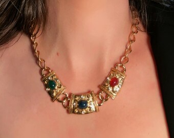 Puede incluir: Un collar de cadena en tono dorado con tres eslabones metálicos rectangulares, cada uno con una piedra cabujón central en verde, azul o rojo. Pequeños cristales transparentes están incrustados alrededor de cada piedra en los eslabones.