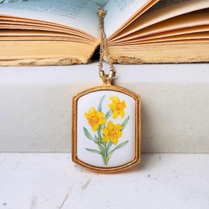 May include: A gold-toned rectangular pendant necklace featuring a white ceramic centre inlay. The inlay displays a detailed illustration of three yellow daffodils with green stems and leaves. The pendant hangs from a delicate gold-toned metal chain.