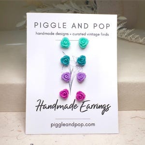 May include: A white card displays handmade rose earrings in teal, purple, and pink. The card reads "Piggle and Pop" and "Handmade Earrings" with the website address. The earrings are arranged in pairs, showcasing their colors and design.