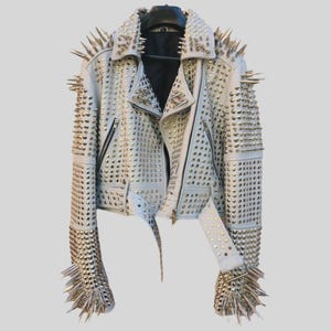 Puede incluir: Una chaqueta de cuero blanca con una extensa variedad de tachuelas metálicas plateadas y púas largas y afiladas que cubren los hombros, mangas, cuello y paneles frontales. La chaqueta incluye un forro interior negro, cremallera y un cinturón.