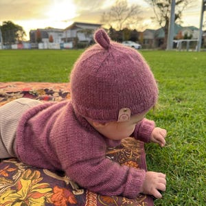May include: A dusty rose knitted long-sleeve jumper and a matching beanie with a small top knot. The beanie features a small, light-coloured rectangular leather tag on the side. The knit fabric has a soft, textured appearance with ribbed cuffs.