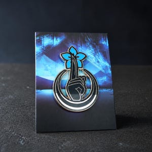 Op de afbeelding: Een metalen emaille speld met een zwarte hand die een stiltegebaar maakt, bekroond met een felblauwe vijfbladige bloem, omlijst door een zwart-witte halve maan, bevestigd op een rechthoekige kaart met een blauw abstract ontwerp.