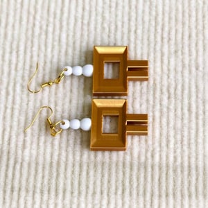 May include: A pair of gold-toned earrings featuring a rectangular frame with a central cutout and three horizontal bars extending from one side. Each earring is attached to a gold hook by a short strand of three small, round, white beads.