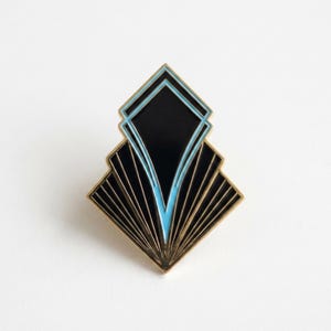 Peut inclure: Une broche émaillée géométrique présentant un centre en forme de losange noir et bleu clair encadré de métal doré. Le design comprend des lignes rayonnantes en éventail partant de la pointe inférieure, créant un aspect symétrique et angulaire.