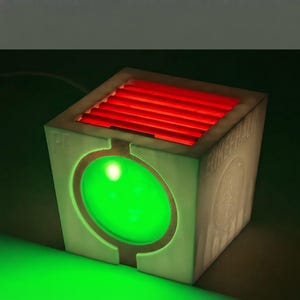 May include: A cube-shaped light fixture with a matte white finish. The front features a circular green glowing light, while the top has a series of horizontal red glowing slats. The side of the cube displays the text ENKEPHALIN and a circular emblem.