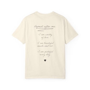Puede incluir: Una camiseta de manga corta de color crema con texto negro en cursiva en la parte posterior que dice: Repeat after me: I am worthy of love. I am beautiful inside and out. I am grateful every day. Un pequeño icono de corazón negro aparece debajo.
