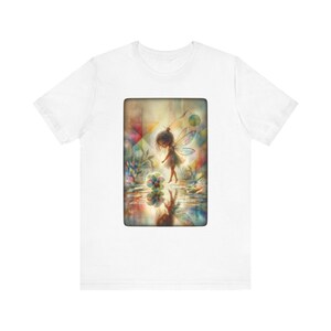 Puede incluir: Una camiseta blanca de manga corta con un estampado gráfico rectangular en el pecho. La imagen muestra una pequeña figura alada junto a una flor, sobre un fondo abstracto y colorido con un reflejo de la escena en el agua.