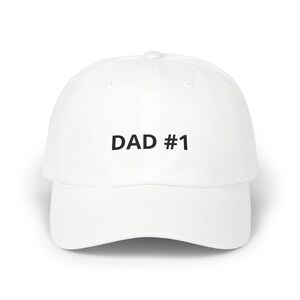 Puede incluir: Una gorra de béisbol blanca con el texto DAD #1 impreso en negro en la parte frontal. La gorra tiene una corona estructurada, una visera curva y un botón en la parte superior central. El diseño es sencillo, limpio y centrado.