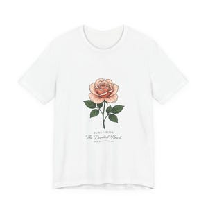 Puede incluir: Una camiseta blanca de manga corta con un gráfico central de una rosa color melocotón con hojas verdes. Debajo de la rosa, el texto dice: JUNE ROSE, The Devoted Heart, fresh flower keepsake. El diseño está centrado en el pecho de la prenda.