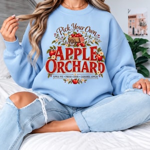 Puede incluir: Una sudadera azul claro de cuello redondo con un estampado gráfico que dice Pick Your Own APPLE ORCHARD, ilustrada con un carro de madera lleno de manzanas rojas, follaje y el texto APPLE PIE, FRESH CIDER, CARAMEL APPLES.