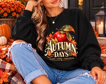 Peut inclure: Un sweat-shirt noir à manches longues orné d'un motif représentant une pomme rouge avec des feuilles vertes et un feuillage d'automne orange. Le texte "AUTUMN DAYS" est écrit dans une police stylisée, avec "SLOW • COZY • GRATEFUL" en dessous.
