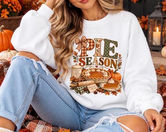 Peut inclure: Un sweat-shirt blanc à manches longues avec une impression graphique portant le texte PIE SEASON. Le motif comprend une tarte à la citrouille, un rouleau à pâtisserie, un bocal et des feuilles d'automne aux tons orange, marron et vert.
