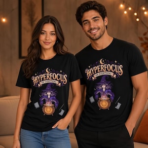 Puede incluir: Una camiseta negra con un estampado gráfico de una bruja con túnica morada detrás de un caldero naranja brillante. Sobre la bruja, el texto "HYPERFOCUS" aparece en una fuente estilizada, con "ON EVERYTHING" escrito en una cinta debajo.