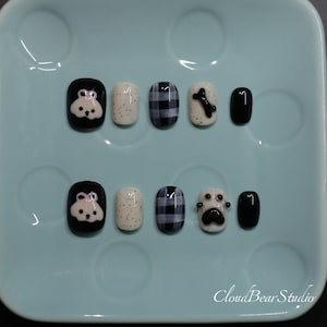May include: A set of ten press-on nails featuring a black and white color palette. Designs include a white dog face on black, cream with black speckles, black and white plaid, a black bone, a black paw print, and solid black nails. CloudBearStudio text.