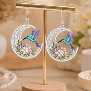 May include: A pair of dangling earrings featuring a white crescent moon shape with embroidered details. Each earring displays a turquoise hummingbird in flight, surrounded by small purple and white flowers with green leaves, hanging from silver hooks.