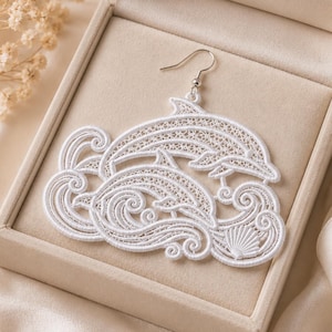 May include: A white lace earring featuring a detailed embroidered design of two dolphins leaping through stylized ocean waves, accented with a small seashell motif at the bottom. The piece is attached to a silver-toned metal hook for hanging.