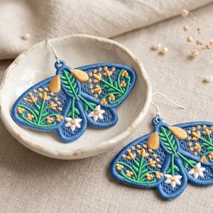 May include: A pair of moth-shaped earrings featuring blue embroidered fabric with yellow and green floral patterns and white flower accents. Each earring is attached to a silver-toned metal hook for hanging, showcasing detailed needlework.