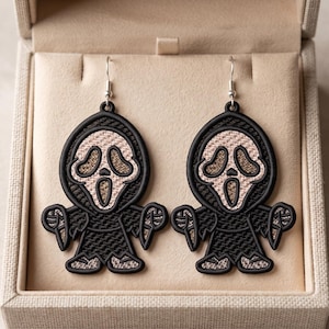 May include: A pair of dangle earrings featuring a black and beige embroidered ghost figure with a hooded cloak and a wide-mouthed expression. The design includes a black outline and silver-toned metal earring hooks for pierced ears.