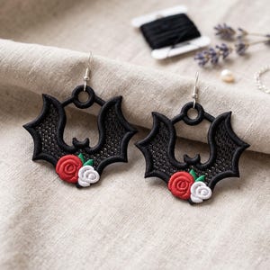 May include: A pair of black embroidered bat-shaped earrings featuring a detailed lace-like wing pattern. Each earring is accented with a small red rose, a white rose, and two green leaves at the bottom, suspended from silver-toned metal earring hooks.