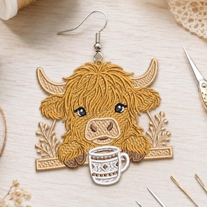 May include: A dangling earring featuring an embroidered golden-brown Highland cow with long shaggy hair and horns, resting its front hooves on a horizontal bar above a small white mug with a brown geometric pattern and a silver hook.