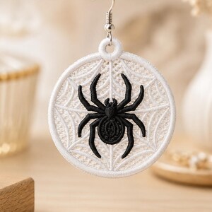May include: A circular embroidered earring featuring a black spider centered on a white web pattern. The design is stitched onto a white fabric disc with a matching white border, suspended from a silver-toned metal hook for ear attachment.