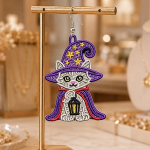 May include: An embroidered earring featuring a white cat wearing a purple witch hat with yellow stars and a purple cape with red trim. The cat is holding a small black lantern with a yellow light. The earring hangs from a silver-toned hook.