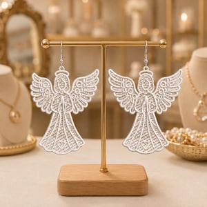 May include: A pair of white lace angel earrings featuring intricate embroidery, a halo, and hands pressed together in prayer. The angel figures have detailed wings and a heart motif at the base of the dress, suspended from silver-toned metal hooks.