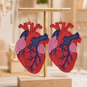 Op de afbeelding: Een paar oorbellen in de vorm van een anatomisch hart met gedetailleerd borduurwerk in rood, donkerblauw en lichtroze. De oorbellen tonen menselijke harten met zichtbare aderen en slagaders, hangend aan zilverkleurige metalen haken.