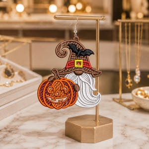 May include: An embroidered earring featuring a gnome wearing a brown witch hat with a black bat and a red band with a yellow buckle. The gnome has a long white beard and is holding a bright orange jack-o'-lantern pumpkin with a carved face.