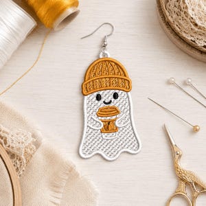 Könnte beinhalten: Ein gestickter Geister-Ohrring, der einen weißen Geist mit einer senfgelben Mütze zeigt, der einen passenden senfgelben Kaffeebecher mit einem kleinen Kürbis-Symbol hält. Der Geist hat schwarze Augen und einen lächelnden Mund an einem Silberhaken.