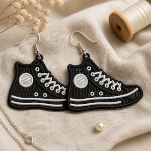 May include: A pair of black and white high-top sneaker earrings featuring detailed embroidery. The design includes white laces, a circular ankle patch, and a white sole. Each earring hangs from a silver-toned metal hook, showcasing a textured fabric finish that mimics the appearance of a classic canvas shoe.