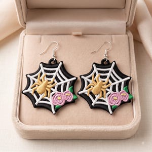 May include: A pair of dangle earrings featuring a black and white embroidered spider web design. Each earring displays a golden yellow spider and two small pink flowers with green leaves. The earrings hang from silver-toned metal hooks.