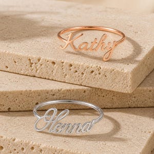 May include: Two thin metal rings with cursive name lettering. The top ring is rose gold-toned and features the name Kathy. The bottom ring is silver-toned and features the name Henna. Both rings have a smooth, polished finish and a delicate band.