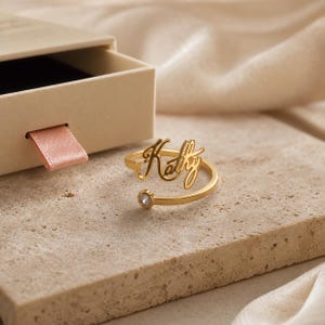 May include: A gold-toned open ring featuring the name Kathy in a cursive script font. The ring has a small, clear, round gemstone set at one end of the band. The piece is displayed next to a beige box with a pink ribbon pull tab.