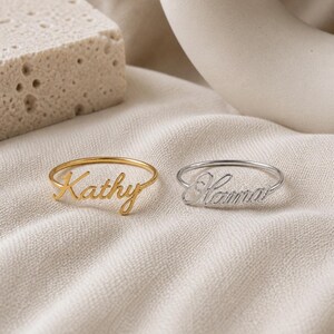 May include: Two thin metal rings featuring custom script lettering. One ring is gold-toned with the name Kathy, and the other is silver-toned with the name Hanna. Both rings have a delicate band design and are displayed side by side on a fabric surface.