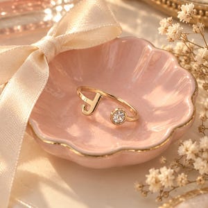 Puede incluir: Un anillo abierto de tono dorado con la letra J en un extremo y una piedra redonda transparente engastada en el otro. El anillo descansa dentro de un pequeño plato de cerámica rosa claro con bordes festoneados, borde dorado y una cinta color crema.