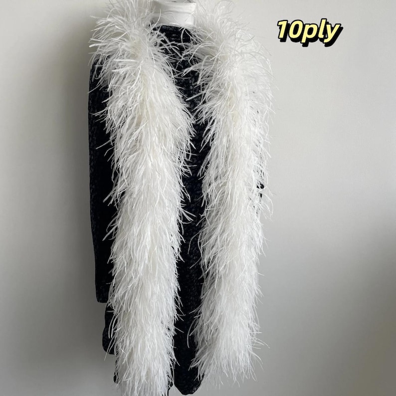 Puede incluir: Una boa de plumas blanca larga con un grosor de 10ply, colocada sobre una prenda negra texturizada. La boa presenta plumas blancas densas y esponjosas que cuelgan verticalmente, creando una apariencia voluminosa sobre la tela oscura.