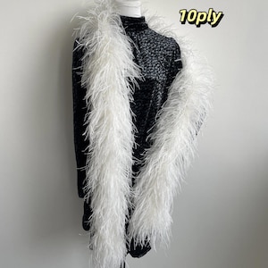 Puede incluir: Una boa de plumas blanca larga con un grosor de 10ply drapeada sobre un vestido negro de manga larga con un sutil patr&oacute;n floral oscuro. La boa consta de numerosas plumas blancas finas y ligeras que crean una apariencia voluminosa y texturizada.