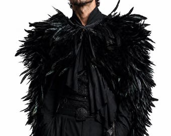 Puede incluir: Una capa de plumas negras sobre una camisa y pantalones negros. La capa presenta plumas negras densas y superpuestas que cubren los hombros y la parte superior de los brazos, atada al cuello con cintas negras finas. El conjunto incluye un cinturón negro.