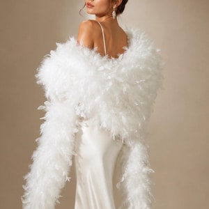 Puede incluir: Una estola de plumas blancas drapeada sobre los hombros, con plumas largas, suaves y esponjosas que crean una textura voluminosa. La estola se lleva sobre un vestido largo de satén blanco, sin mangas, con tirantes finos.