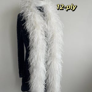 Puede incluir: Una boa de plumas blanca larga con un grosor de 12 capas, colocada sobre una prenda oscura de manga larga con estampado. El texto 12-ply es claramente visible en la esquina superior derecha de la imagen, indicando la densidad de las plumas.