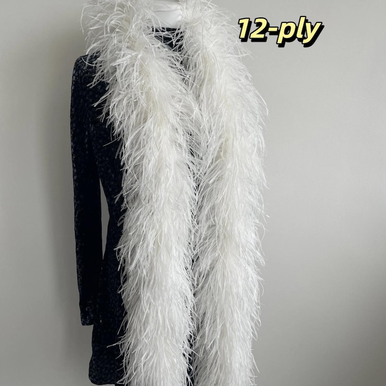 Puede incluir: Una boa de plumas blanca larga con un grosor de 12 capas, colocada sobre una prenda oscura de manga larga con estampado. El texto 12-ply es claramente visible en la esquina superior derecha de la imagen, indicando la densidad de las plumas.
