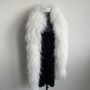 Puede incluir: Una boa larga de plumas de avestruz blancas drapeada sobre un vestido negro sin mangas con un sutil estampado floral en tonos oscuros. La boa presenta plumas blancas densas y esponjosas que se extienden desde el cuello hasta el dobladillo.