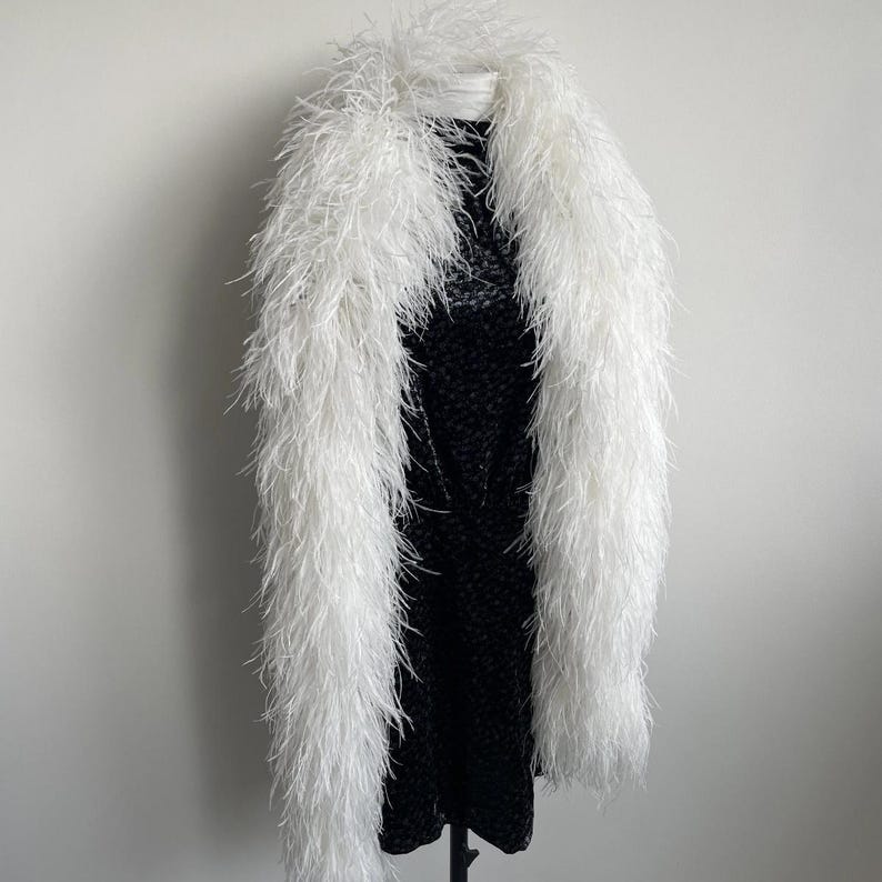 Puede incluir: Una boa larga de plumas de avestruz blancas drapeada sobre un vestido negro sin mangas con un sutil estampado floral en tonos oscuros. La boa presenta plumas blancas densas y esponjosas que se extienden desde el cuello hasta el dobladillo.