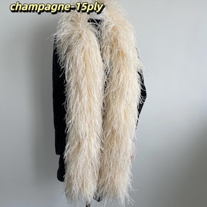 Puede incluir: Un boa de plumas largo de color champ&aacute;n con una construcci&oacute;n de 15 capas, drapeado sobre una prenda negra. El texto 'champagne-15ply' es visible en la esquina superior izquierda. Las plumas son largas, vaporosas y de color beige claro.