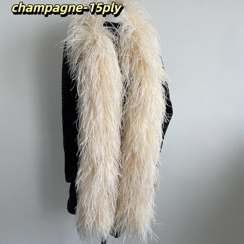 Puede incluir: Un boa de plumas largo de color champ&aacute;n con una construcci&oacute;n de 15 capas, drapeado sobre una prenda negra. El texto 'champagne-15ply' es visible en la esquina superior izquierda. Las plumas son largas, vaporosas y de color beige claro.
