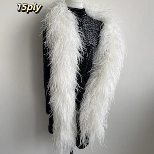 Puede incluir: Una boa de plumas blancas de 15ply drapeada sobre una prenda superior negra de manga larga con un sutil patr&oacute;n floral oscuro. La boa presenta plumas blancas largas y tenues que crean una textura voluminosa, contrastando con la prenda oscura.